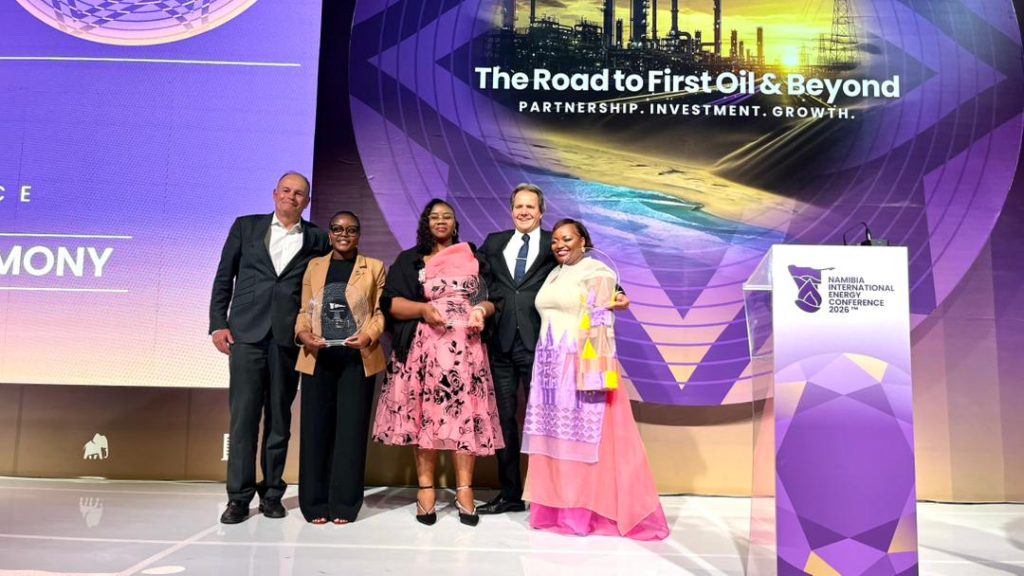 WhatsApp Image 2026-04-16 at 110904 AM | Mining and Energy Namibia | Namibia’s Leading Mining & Energy News Five people stand on a stage posing with a glass trophy at the Namibia International Energy Conference 2026, with a purple backdrop reading 'The Road to First Oil & Beyond.'