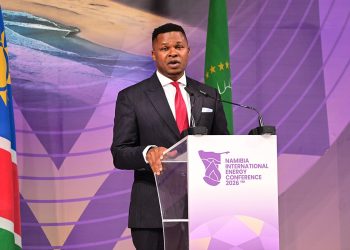 Male speaker in a dark suit and red tie delivers a speech at a podium labeled Namibia International Energy Conference 2026, with flags in the background.