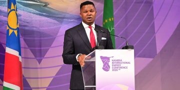 Male speaker in a dark suit and red tie delivers a speech at a podium labeled Namibia International Energy Conference 2026, with flags in the background.