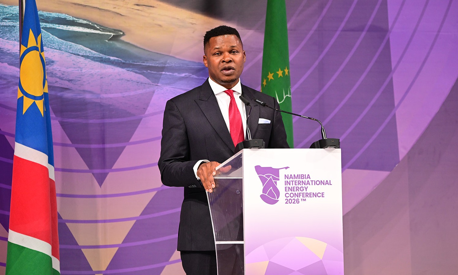 Male speaker in a dark suit and red tie delivers a speech at a podium labeled Namibia International Energy Conference 2026, with flags in the background.