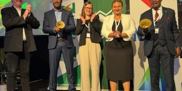 Five professionals on a stage at an awards event, two men holding gold trophies while others applaud, backdrop banners visible.