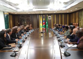 A large oval conference table with many people seated on both sides in formal attire, flags visible in the background.