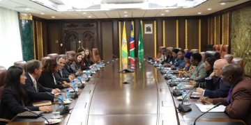 A large oval conference table with many people seated on both sides in formal attire, flags visible in the background.