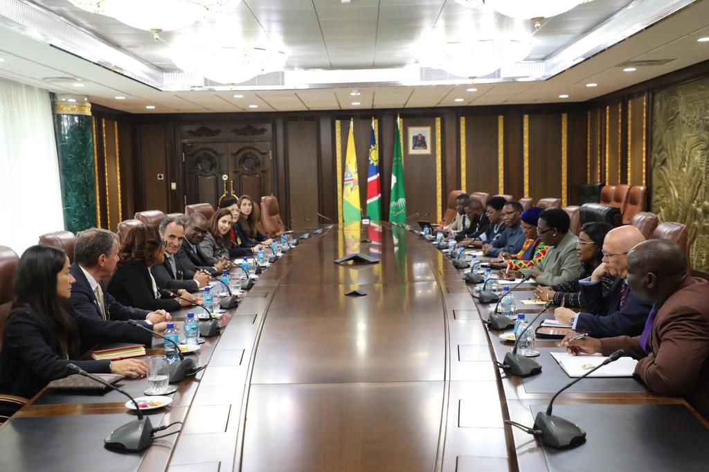 A large oval conference table with many people seated on both sides in formal attire, flags visible in the background.