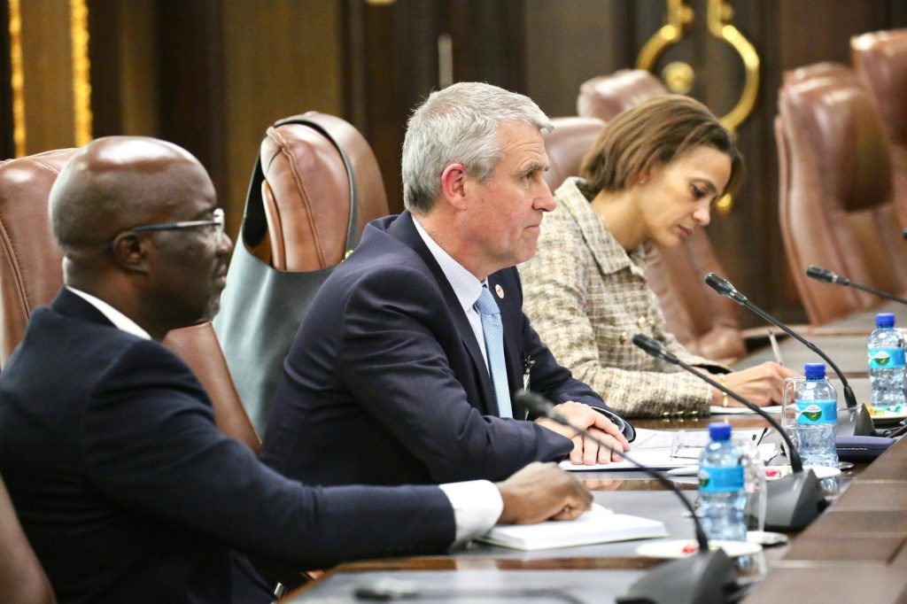 Three professionals sit at a long conference table with microphones, taking notes during a meeting.