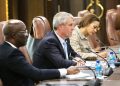 Three professionals sit at a long conference table with microphones, taking notes during a meeting.