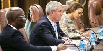 Three professionals sit at a long conference table with microphones, taking notes during a meeting.