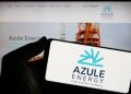 Close-up of a hand holding a white card with the Azule Energy logo in blue, blurred Azule Energy website on a screen in the background.