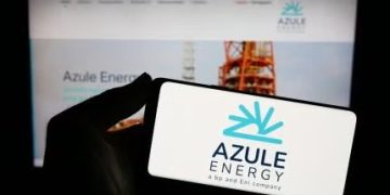 Close-up of a hand holding a white card with the Azule Energy logo in blue, blurred Azule Energy website on a screen in the background.