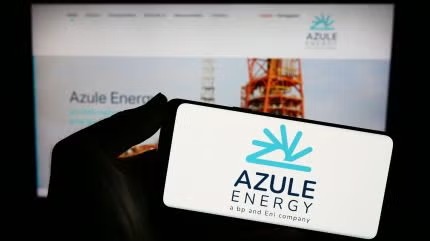 Close-up of a hand holding a white card with the Azule Energy logo in blue, blurred Azule Energy website on a screen in the background.