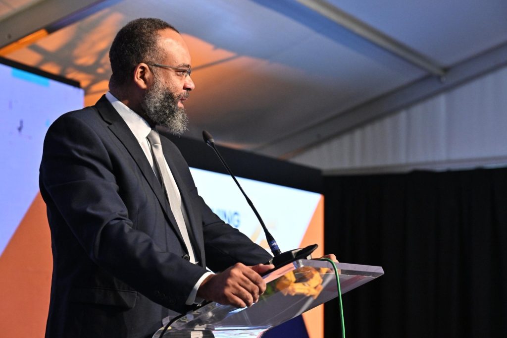 Bearded man in a dark suit and tie delivering a speech at a podium with a microphone at a conference stage.