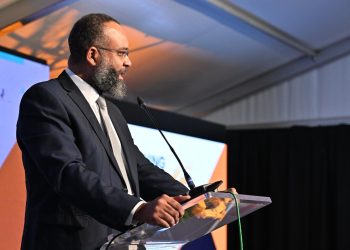 Bearded man in a dark suit and tie delivering a speech at a podium with a microphone at a conference stage.