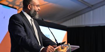 Bearded man in a dark suit and tie delivering a speech at a podium with a microphone at a conference stage.