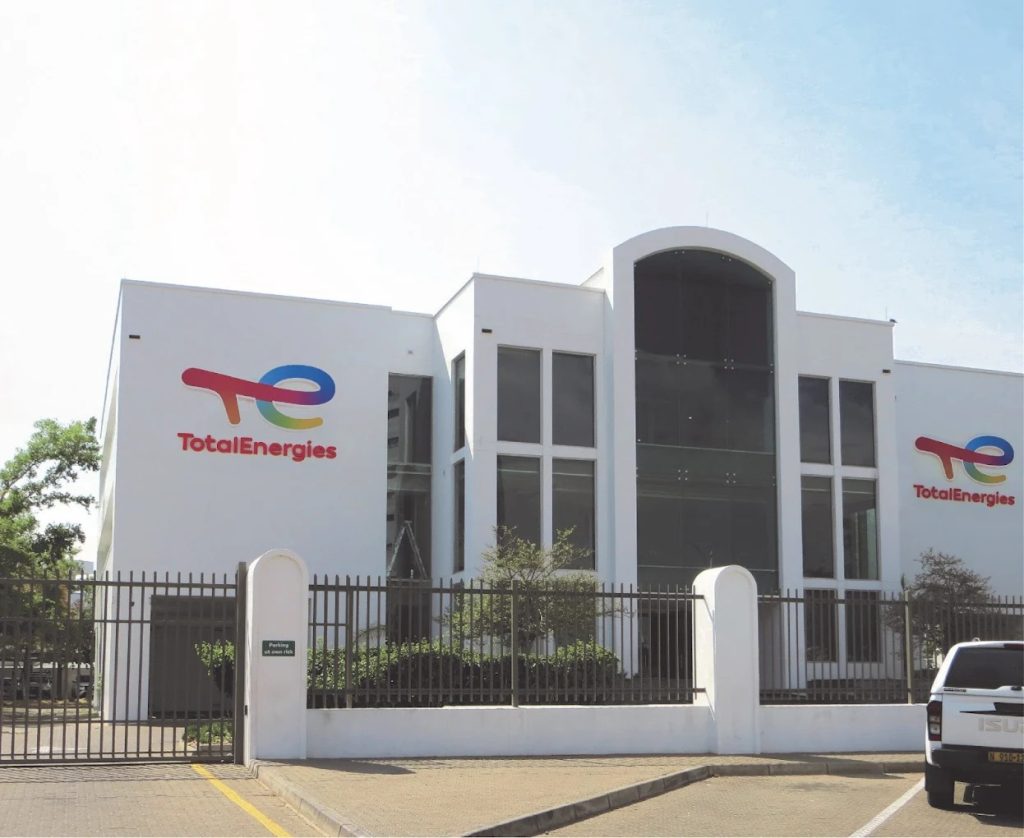 White TotalEnergies office building with large central glass entrance, logo on left and right walls, gated front yard, and a white pickup parked nearby.