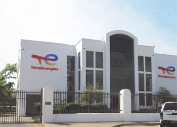 White TotalEnergies office building with large central glass entrance, logo on left and right walls, gated front yard, and a white pickup parked nearby.