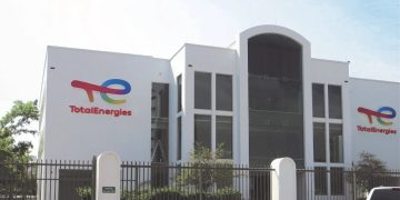 White TotalEnergies office building with large central glass entrance, logo on left and right walls, gated front yard, and a white pickup parked nearby.