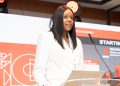 Woman in a white blazer speaks at a podium during a conference, standing behind a clear lectern with a microphone and red stage backdrop.