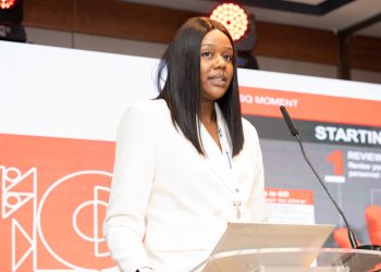 Woman in a white blazer speaks at a podium during a conference, standing behind a clear lectern with a microphone and red stage backdrop.