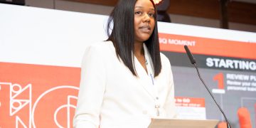 Woman in a white blazer speaks at a podium during a conference, standing behind a clear lectern with a microphone and red stage backdrop.