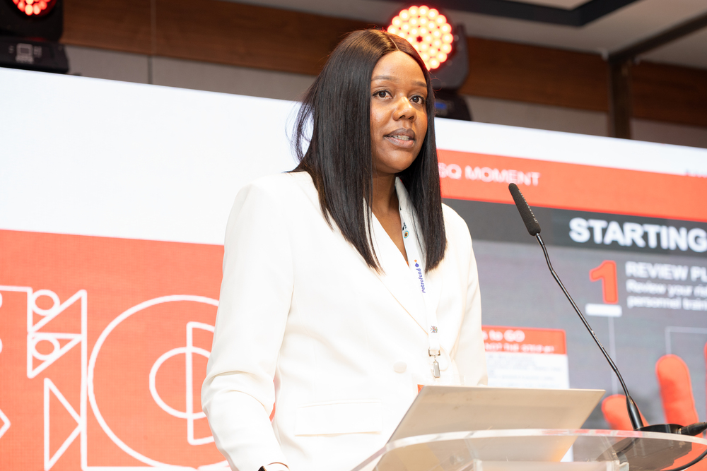 Woman in a white blazer speaks at a podium during a conference, standing behind a clear lectern with a microphone and red stage backdrop.