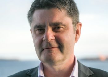 Portrait of a man in a dark blazer and light pink shirt, smiling outdoors with water in the background.