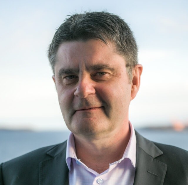 Portrait of a man in a dark blazer and light pink shirt, smiling outdoors with water in the background.