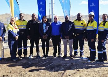 Group of professionals in safety vests posing on a dirt construction site with blue flags in the background.