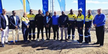 Group of professionals in safety vests posing on a dirt construction site with blue flags in the background.