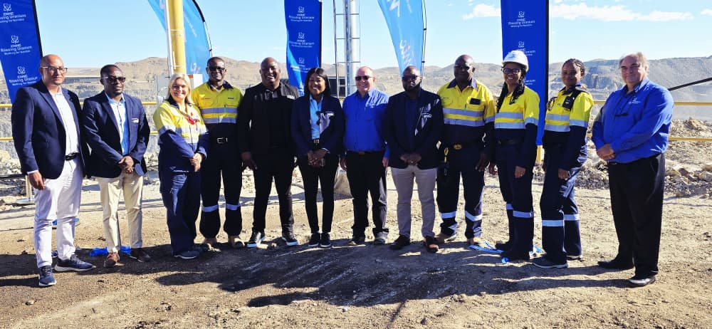 Group of professionals in safety vests posing on a dirt construction site with blue flags in the background.