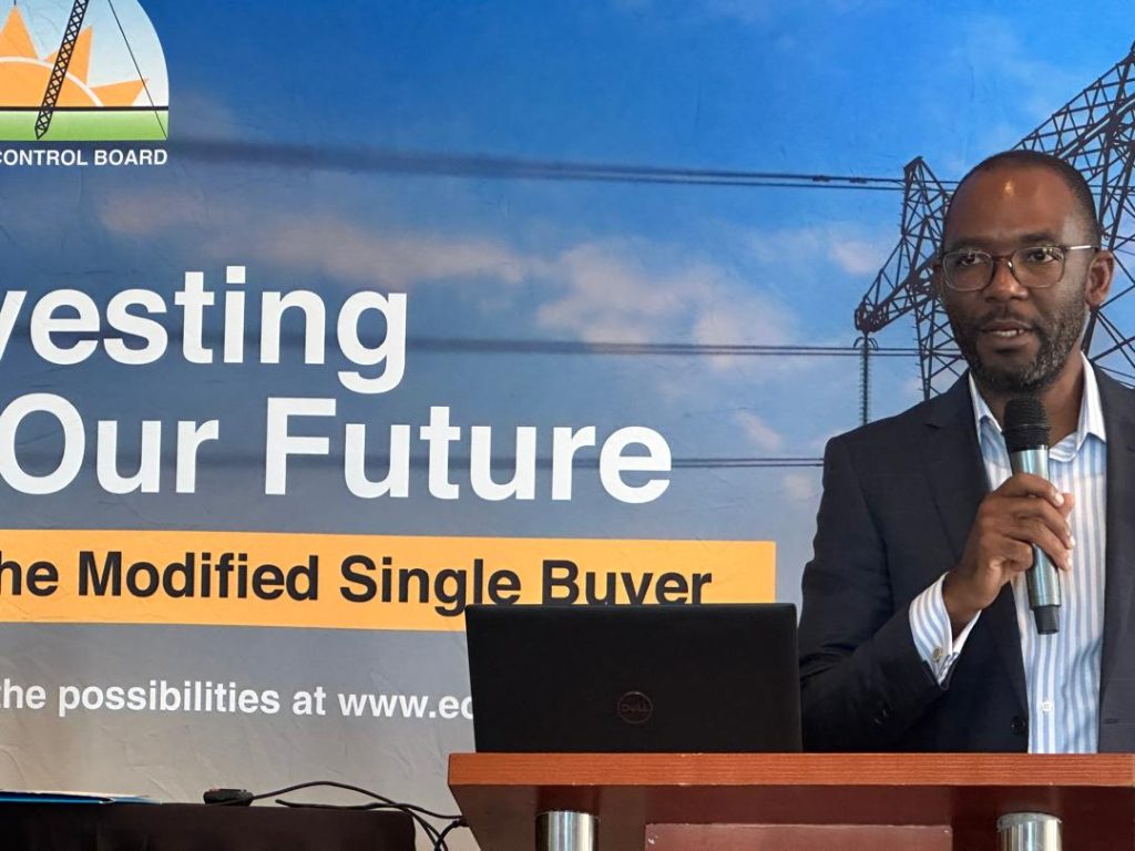 Man in a suit speaks into a handheld microphone at a podium in front of a banner about investing our future and a modified single buyer program.