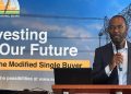 Man in a suit speaks into a handheld microphone at a podium in front of a banner about investing our future and a modified single buyer program.