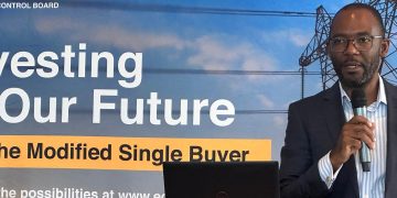 Man in a suit speaks into a handheld microphone at a podium in front of a banner about investing our future and a modified single buyer program.