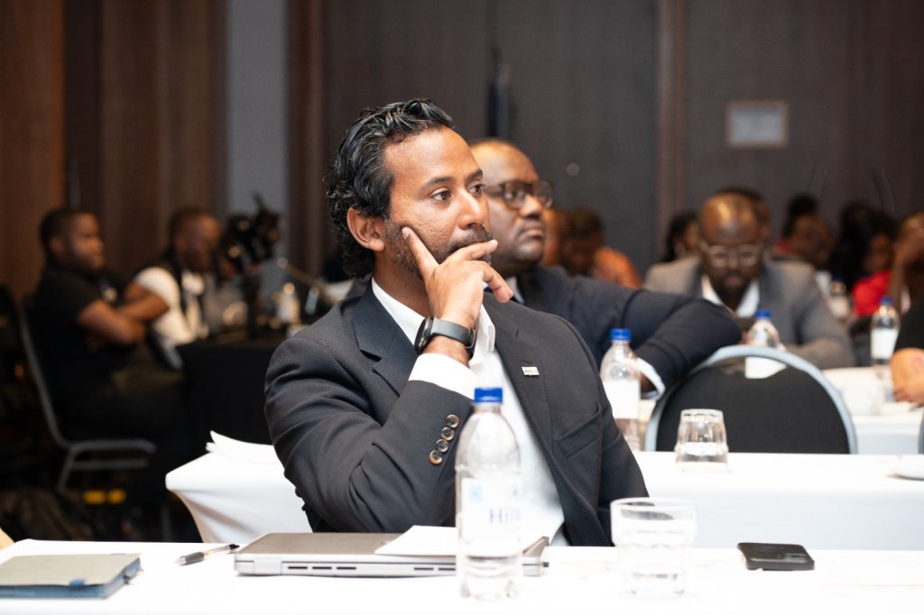 Business professional in a dark suit sits at a conference table, resting his chin on his hand in thought while others listen in the background.