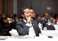 Business professional in a dark suit sits at a conference table, resting his chin on his hand in thought while others listen in the background.