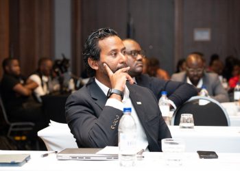 Business professional in a dark suit sits at a conference table, resting his chin on his hand in thought while others listen in the background.