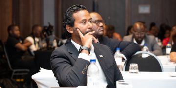 Business professional in a dark suit sits at a conference table, resting his chin on his hand in thought while others listen in the background.