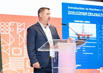 Man in a dark suit giving a presentation at a clear acrylic podium, with a large blue slide behind him.