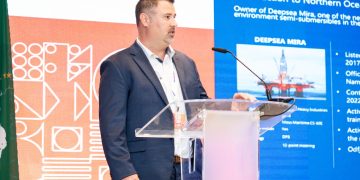 Man in a dark suit giving a presentation at a clear acrylic podium, with a large blue slide behind him.