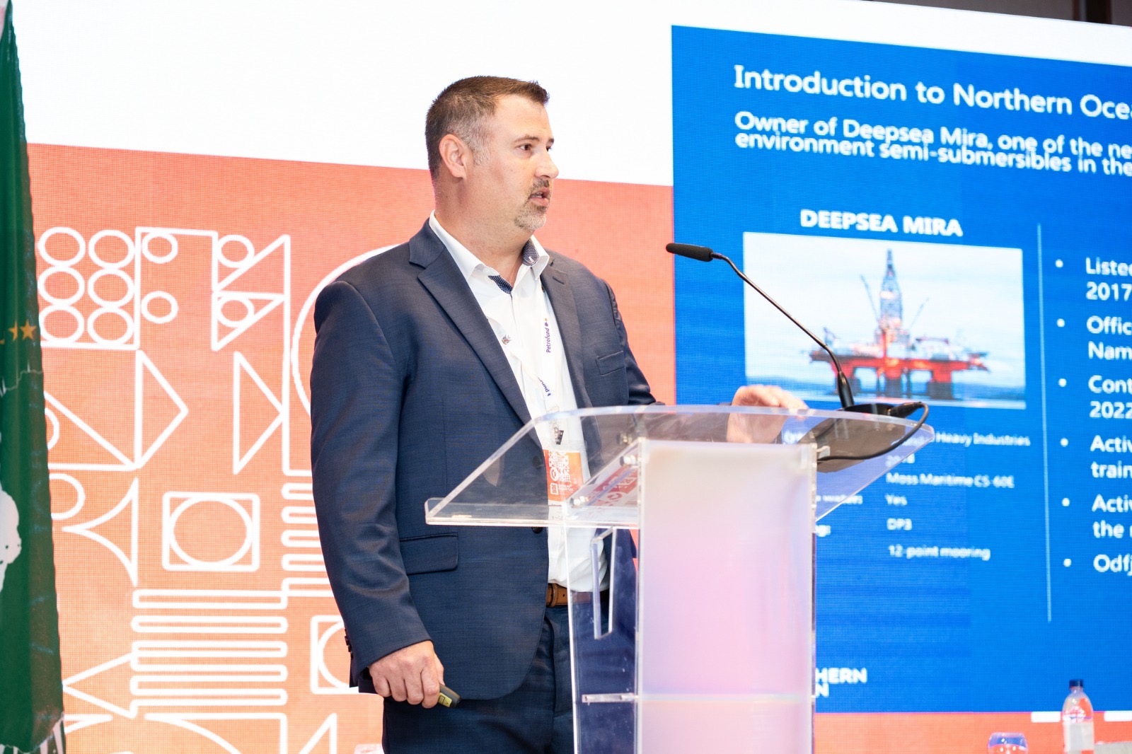 Man in a dark suit giving a presentation at a clear acrylic podium, with a large blue slide behind him.