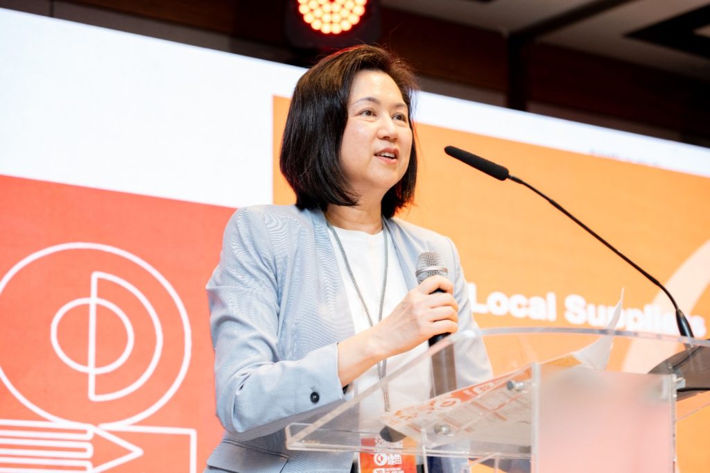 Woman in a light blazer speaking at a podium with a microphone, orange backdrop behind her.