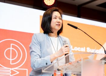 Woman in a light blazer speaking at a podium with a microphone, orange backdrop behind her.