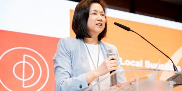 Woman in a light blazer speaking at a podium with a microphone, orange backdrop behind her.