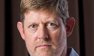 Close-up portrait of a middle-aged man with light brown hair, blue eyes, and stubble, wearing a light blue shirt.