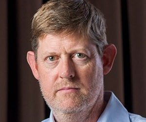 Close-up portrait of a middle-aged man with light brown hair, blue eyes, and stubble, wearing a light blue shirt.