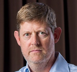 Close-up portrait of a middle-aged man with light brown hair, blue eyes, and stubble, wearing a light blue shirt.