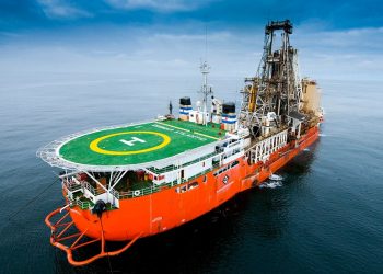 Orange offshore supply vessel with a green helipad on deck and a large drilling rig at the stern, sailing on calm water under a cloudy sky.