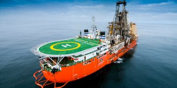 Orange offshore supply vessel with a green helipad on deck and a large drilling rig at the stern, sailing on calm water under a cloudy sky.