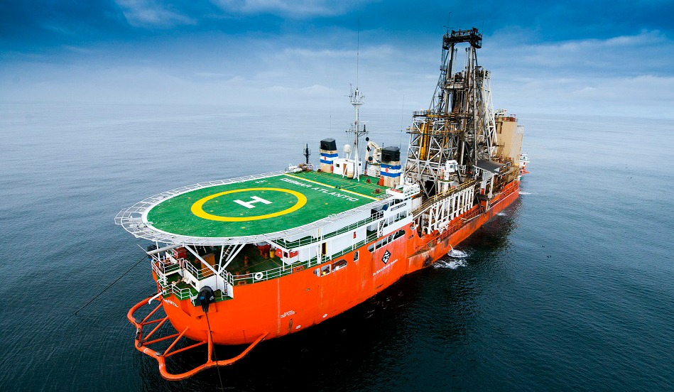 Orange offshore supply vessel with a green helipad on deck and a large drilling rig at the stern, sailing on calm water under a cloudy sky.