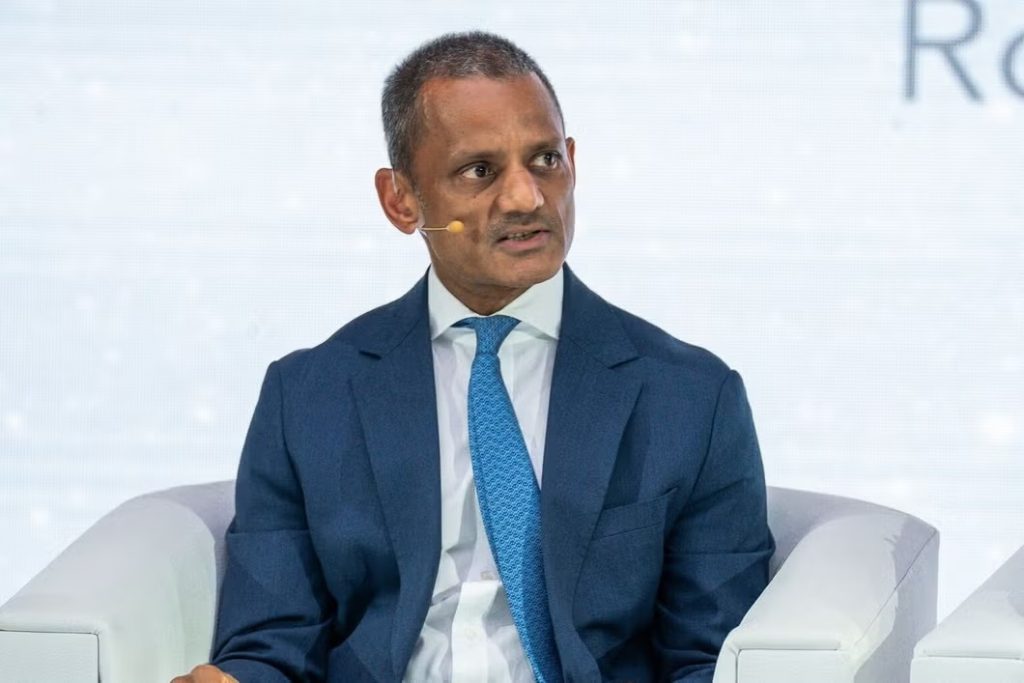 Man in a dark blue suit and light blue tie sits on a stage with a headset microphone, speaking at a panel discussion.