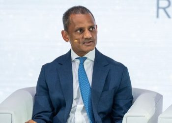Man in a dark blue suit and light blue tie sits on a stage with a headset microphone, speaking at a panel discussion.
